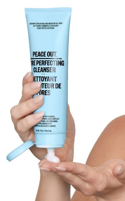 A hand dispensing Peace Out Pore Perfecting Cleanser into the palm. The packaging highlights its creamy exfoliating formula, suitable for all skin types