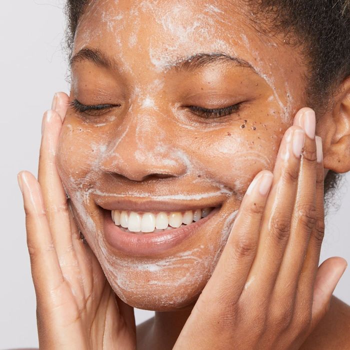 A model smiling, eyes closed, gently massaging their face with foamy cleanser while promoting the skincare product in a shopping article.