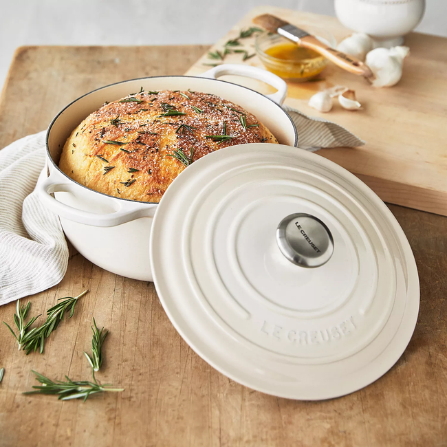 A Le Creuset Dutch oven with a freshly baked loaf of bread inside, resting on a wooden surface, surrounded by sprigs of rosemary and garlic