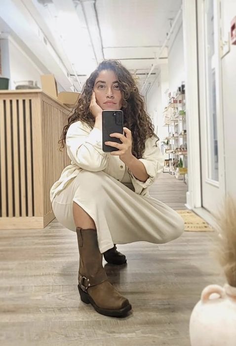 A woman in a white outfit and brown boots taking a mirror selfie while squatting in a room filled with shelves and a counter.