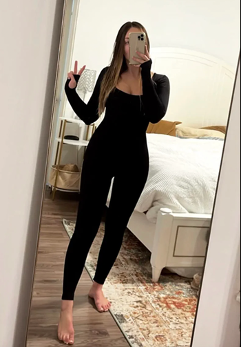 A reviewer is modeling a sleek, black, form-fitting bodysuit that highlights its flattering cut and stretch.