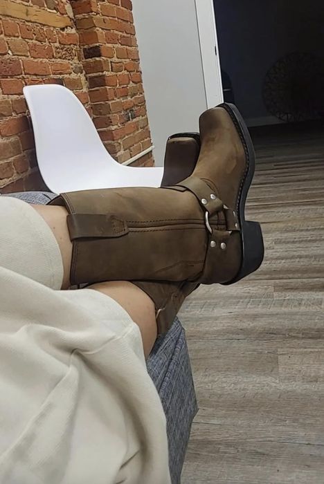 A person lounging with their legs up, wearing tan boots, set against a white chair and a brick wall backdrop.