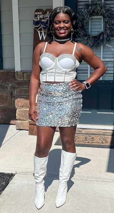 A person stands outside a house in a pearl-studded corset top, a glittering sequin skirt, and white knee-high boots.