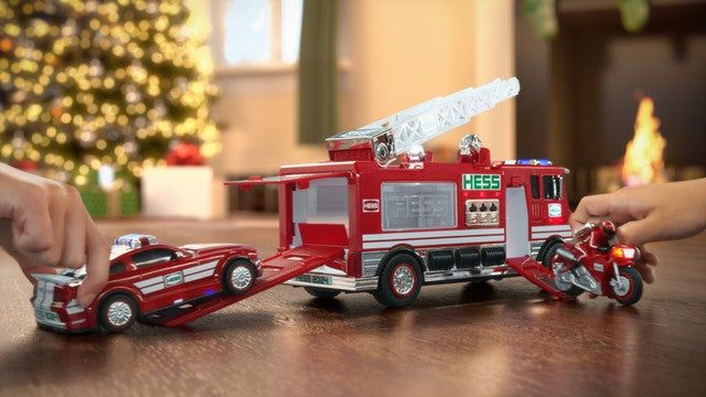 Children playing with a toy set that includes a fire truck, a police car, and a motorcycle on a wooden floor, with a Christmas tree in the background.