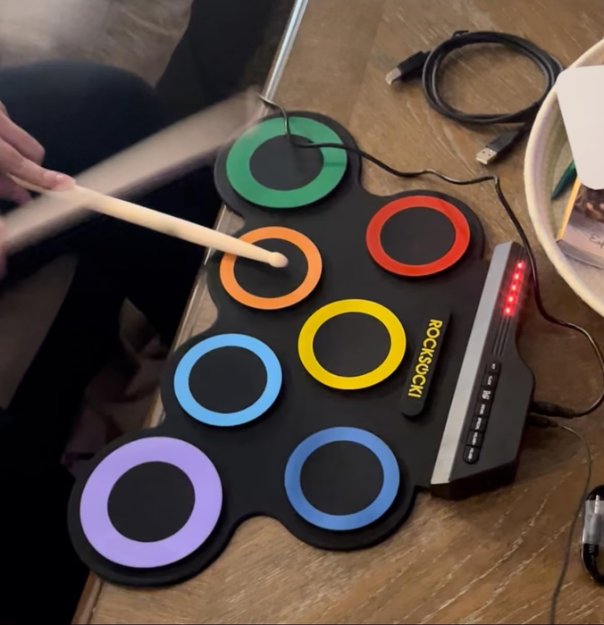 An electronic drum pad with vibrant circles, connected to a device. A person is tapping away with drumsticks on the colorful, circular pad, ideal for practicing music.