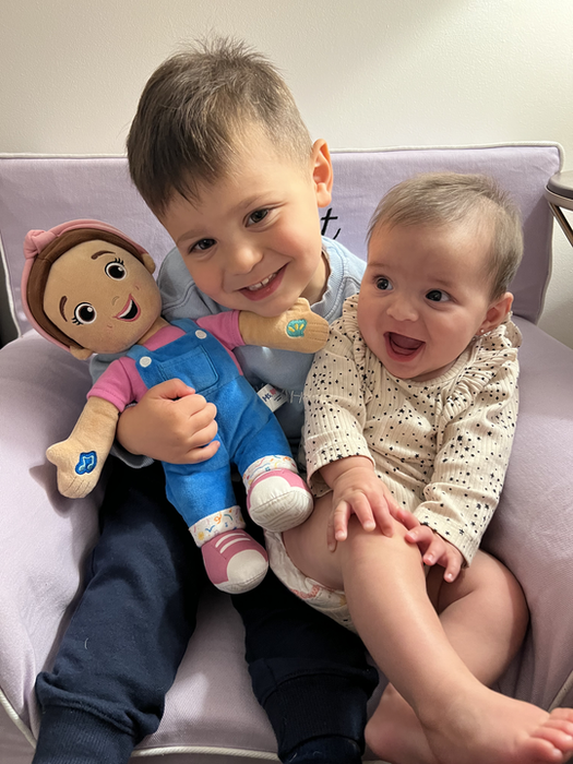 Two joyful children, one holding a plush doll, share a seat together on a cozy chair.