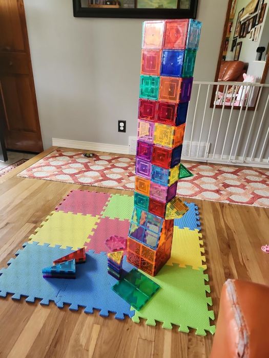 A tall tower built with colorful magnetic tiles placed on a foam mat in a cozy living room, with additional tiles scattered nearby. A kid-friendly play zone is visible.