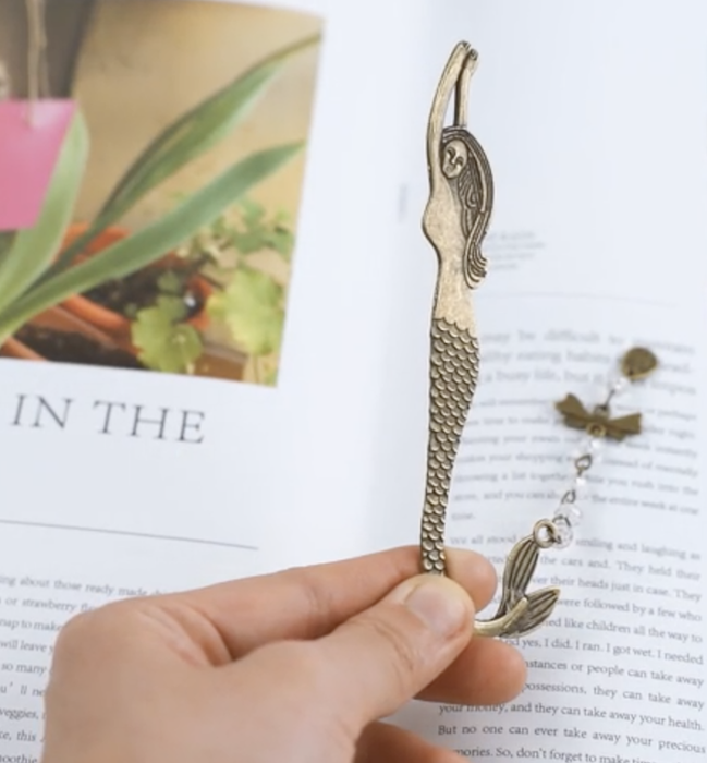 a golden bookmark in the shape of a mermaid with flowing hair. The mermaid's tail is adorned with silver and gold jewels, curving to hang over the page of a book.