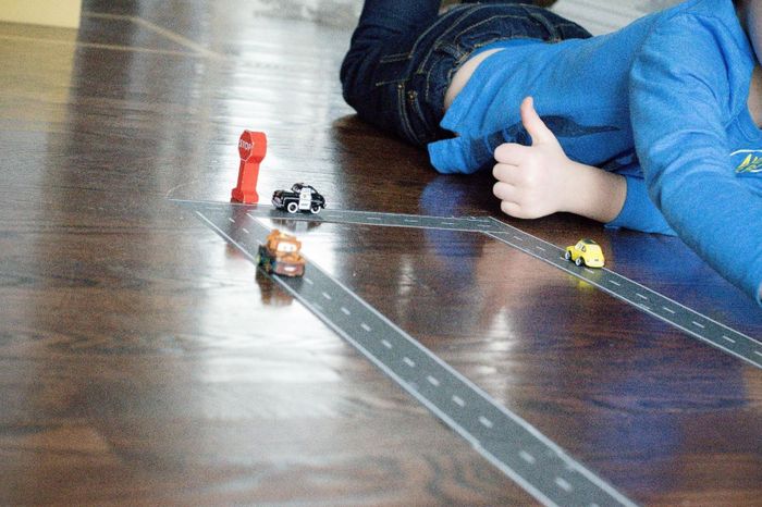 A child playing with toy cars on a tape road layout on the wooden floor, flashing a thumbs-up in excitement