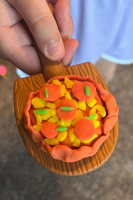 A Play-Doh pizza model with orange and yellow toppings on a small wooden paddle, held by a hand