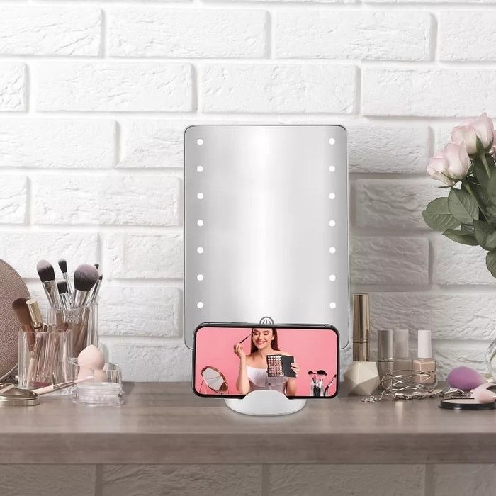 A makeup vanity setup featuring a well-lit mirror, a variety of makeup products, and brushes arranged on the table, with a smartphone showing a woman giving a makeup tutorial.