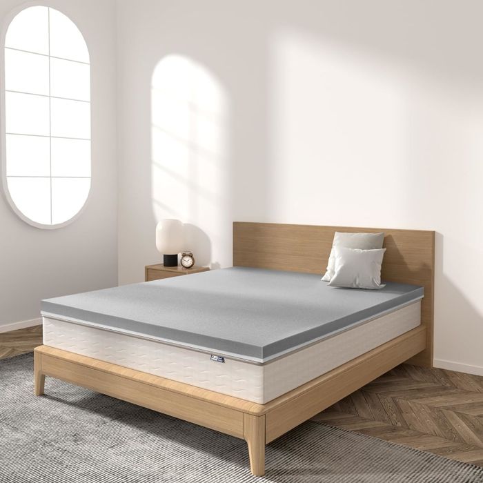 A minimalist bedroom with a well-made bed featuring a gray mattress topper and pillow on a wooden bed frame, accompanied by a lamp and a window.