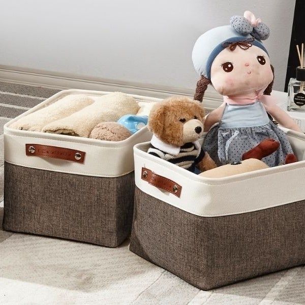 Three fabric storage bins, filled with towels and toys including a teddy bear and a doll, are arranged on a carpeted floor beside a sofa.