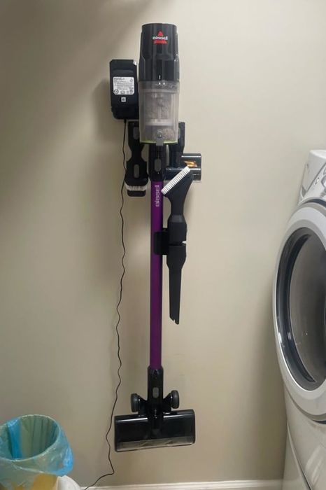 A cordless vacuum cleaner mounted on the laundry room wall next to the washing machine, with various cleaning attachments clearly visible.