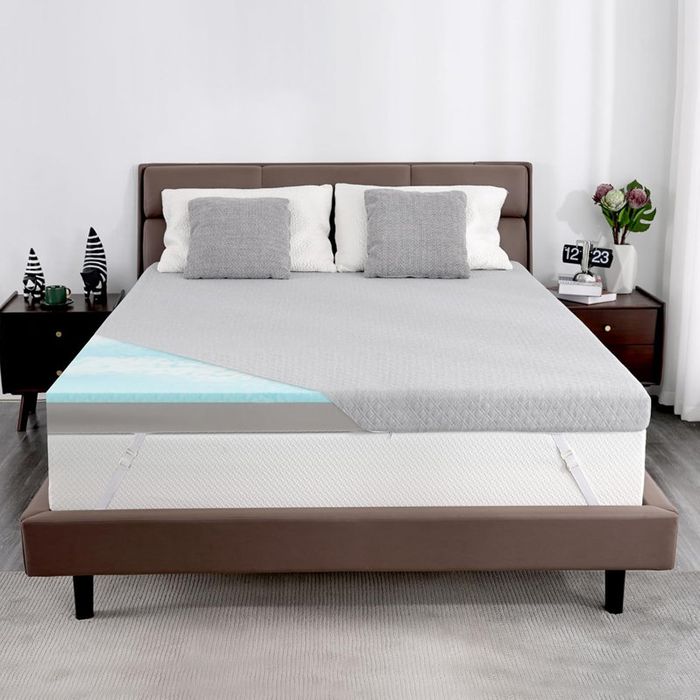 A bed featuring a gray mattress topper in a contemporary bedroom, complemented by decorative pillows, bedside plants, and a calendar on the nightstand.