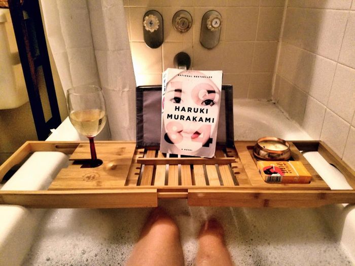 A reviewer's perspective of a wooden bath caddy holding a book and a glass of wine as they relax in a bubble-filled tub