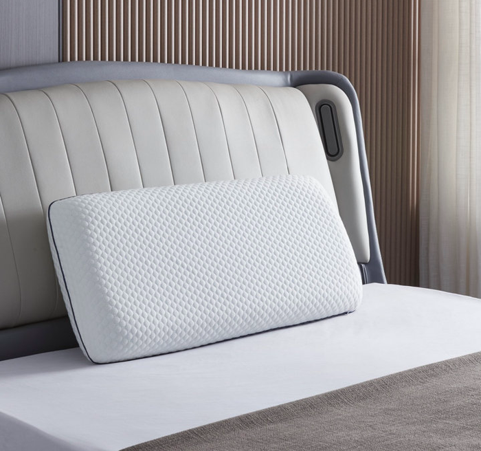 A pillow with a textured cover, placed on a neatly made bed in a contemporary bedroom featuring a padded headboard and sheer curtains in the background