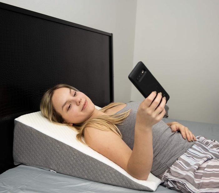 An individual dressed in a gray tank top and striped pajama bottoms is comfortably reclining on a wedge pillow, engaging with a smartphone while relaxing in bed.