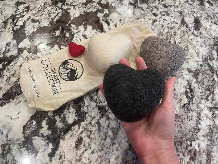Hand holding a wool dryer ball shaped like a heart, surrounded by more dryer balls and a Shanti Collection bag on a marble surface