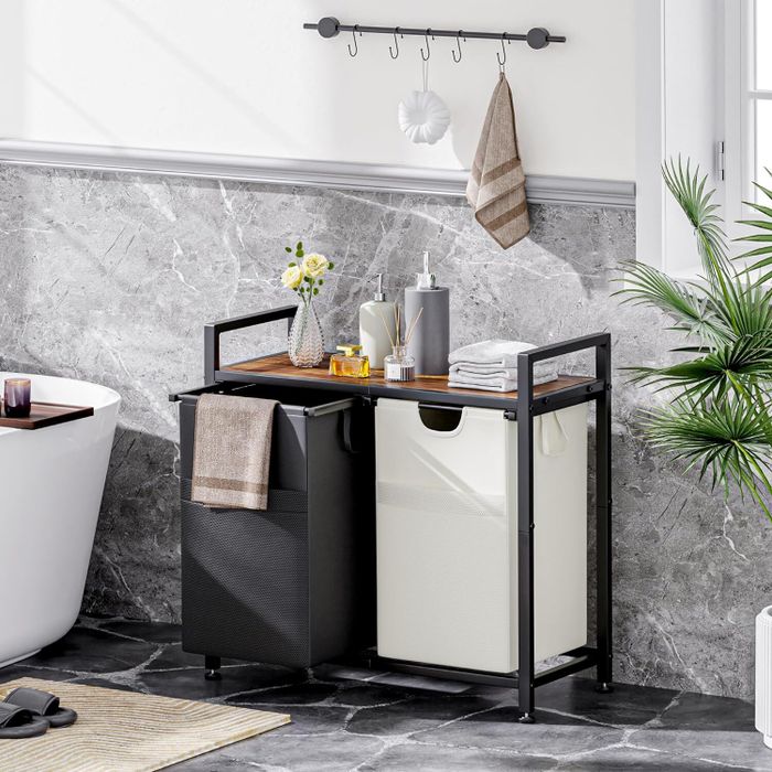 A contemporary bathroom scene showcasing a dual-compartment laundry hamper with a wooden surface holding toiletries and towels, situated near a bathtub and a potted palm plant