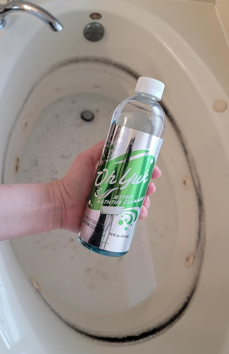 A hand holds a bottle of jetted tub cleaner over a bathtub with a dark grime ring around its edge