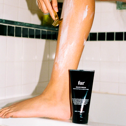 A different model shaving their leg with the sleek cream bottle in front of them. The cream is smooth, not foamy.