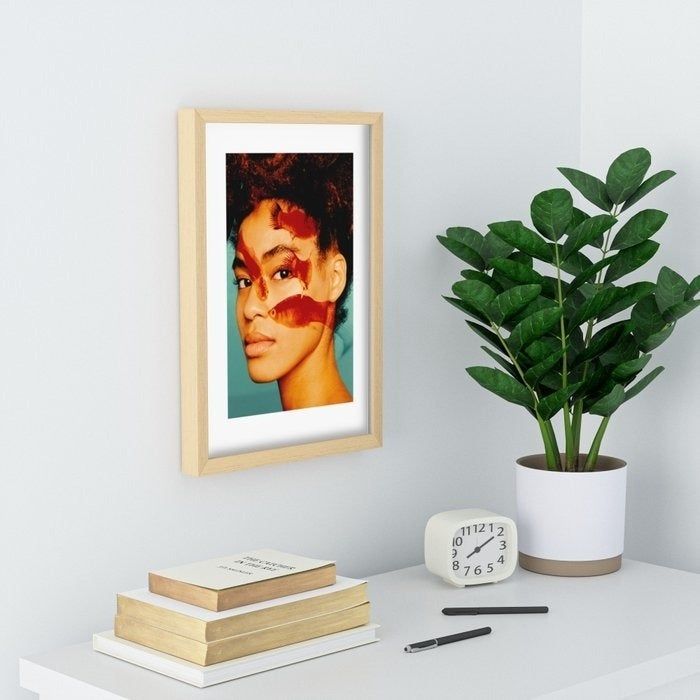 A framed portrait of an enigmatic woman hanging on a wall beside a potted plant and a shelf filled with books