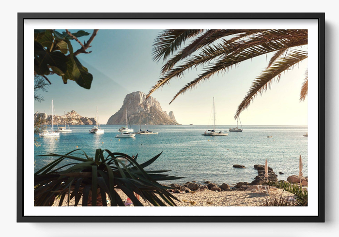 A serene bay view featuring yachts and a rocky outcrop in the distance, framed by palm leaves