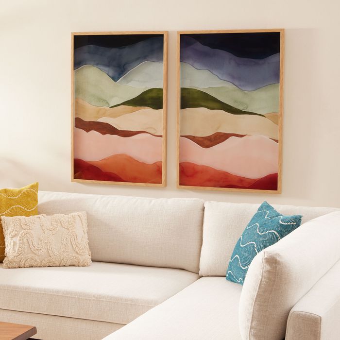 Two contemporary landscape paintings displayed above a beige sectional sofa with decorative pillows, featured in a home decor shopping article