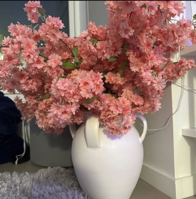 A vibrant bouquet of pink flowers in a white vase, displayed on a shelf alongside books and a carpeted floor