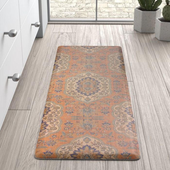 A decorative kitchen mat with a detailed floral and geometric design placed on wooden flooring next to white cabinets, with potted plants by a window.