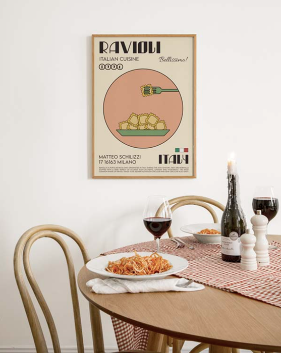 A Ravioli poster hanging above a dining table set with a plate of pasta, wine glasses, and a candle, evoking an Italian-inspired dining atmosphere