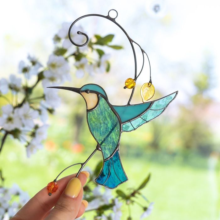 A hand holding a stained glass hummingbird ornament against a floral backdrop, ideal for home decor enthusiasts