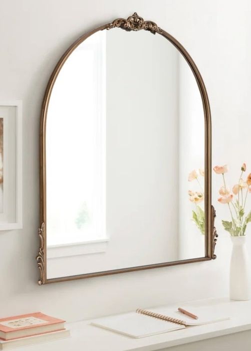A mirror hanging gracefully above a white desk, accompanied by a notebook, pen, and a vase of fresh flowers