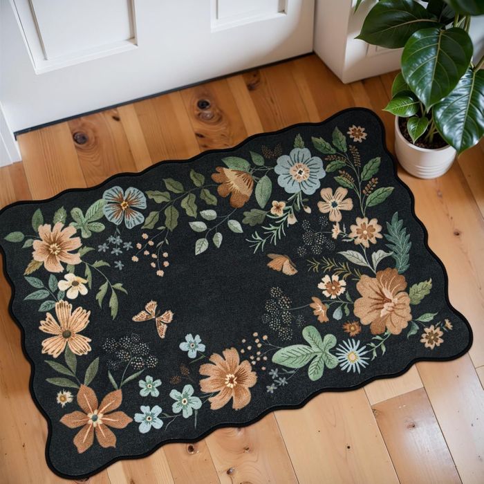 A floral indoor rug with scalloped edges, featuring detailed flower and butterfly embroidery, placed on a wooden floor near a white door, beside a green potted plant.