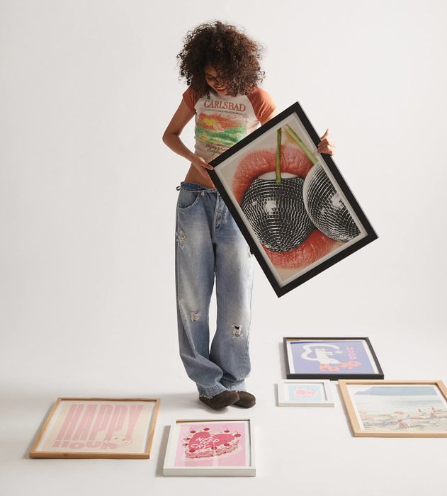 A person holding a framed cherry print, surrounded by various art prints laid out on the floor, dressed in casual jeans and a graphic T-shirt
