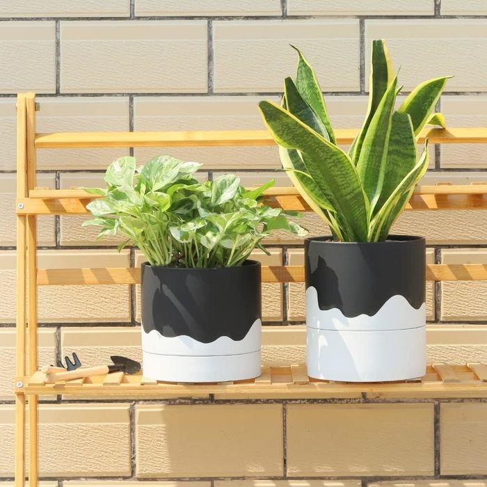 Two black and white planters with dripping paint designs, sitting on a wooden shelf with green plants inside. The planters are placed against a brick wall.