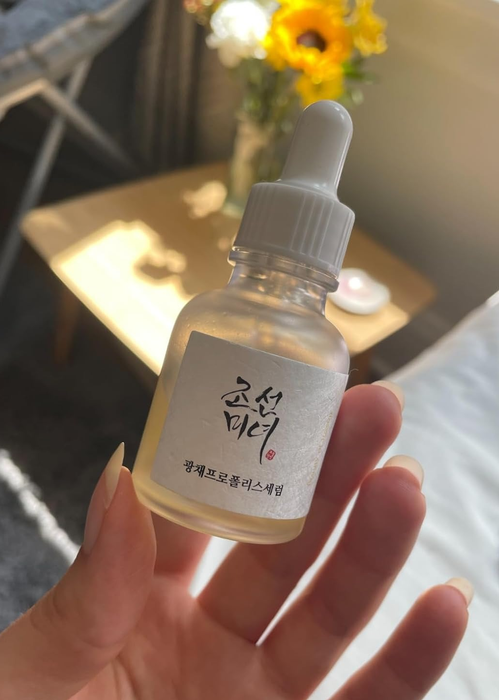 A reviewer holding a dropper bottle of skincare serum near a window, with flowers and a book on the table in the background