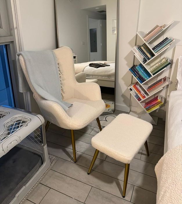 A modern-style chair and ottoman with gold legs placed in a cozy reading nook next to a full bookshelf.