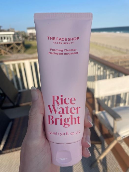 A reviewer holding a tube of The Face Shop Rice Water Bright Foaming Cleanser while standing on a balcony with a beach view