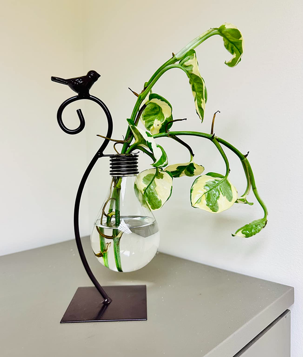 A decorative plant displayed in a lightbulb vase with a metal stand, complete with a bird ornament, resting on a table.
