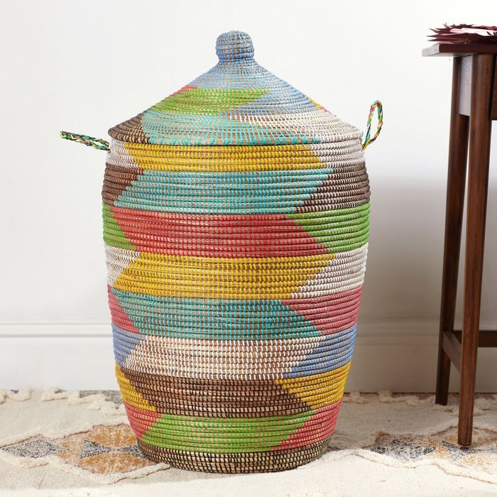 A multicolored woven basket with a lid and side handles, placed on a sandy floor against a white wall, highlighting the exquisite craftsmanship.
