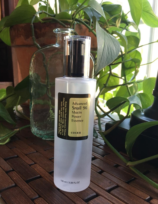 A clear bottle of COSRX Advanced Snail 96 Mucin Power Essence sits on a wooden surface, surrounded by a plant and a glass jar in the backdrop.