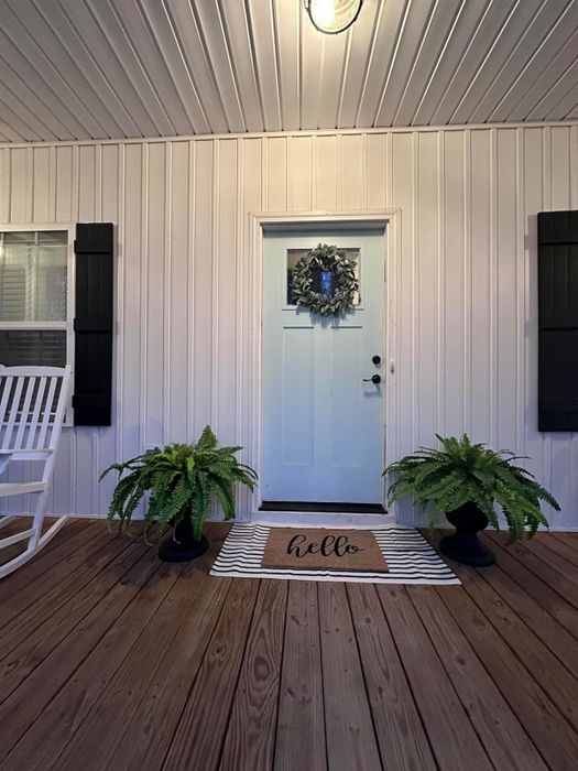 A white front door with a welcoming 'hello' doormat, surrounded by green plants and a festive wreath hanging on the door