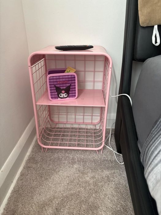 A pink metal storage rack with two shelves, one featuring a small purple basket holding a remote control