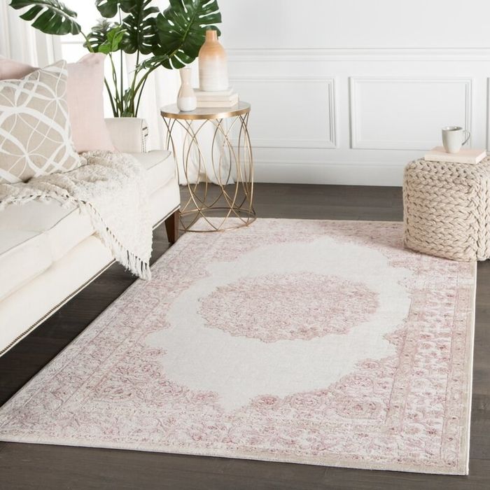 A pink and white medallion area rug set in a living room with a sofa, side table, floor plant, and a woven pouf.