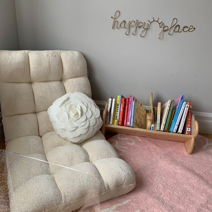 A cozy reading nook featuring a plush chair, a decorative cushion, a wooden 'happy place' sign, and a rack filled with children's books