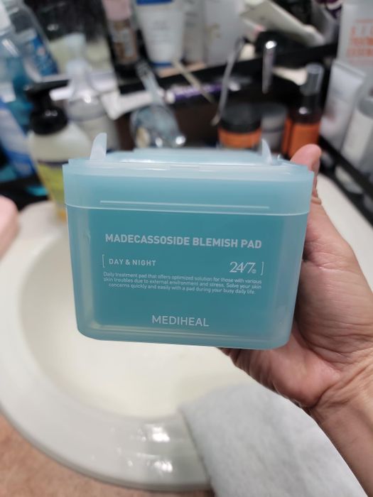 Hand holding a Mediheal Madecassoside Blemish Pad container with skincare products in the background.