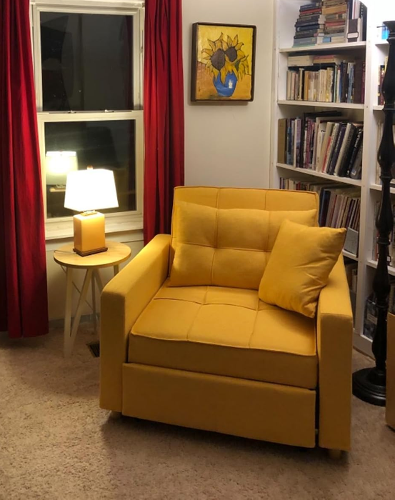 A reviewer's cozy reading corner with a yellow armchair, a side table topped with a lamp, shelves filled with books, and a cheerful sunflower painting on the wall.