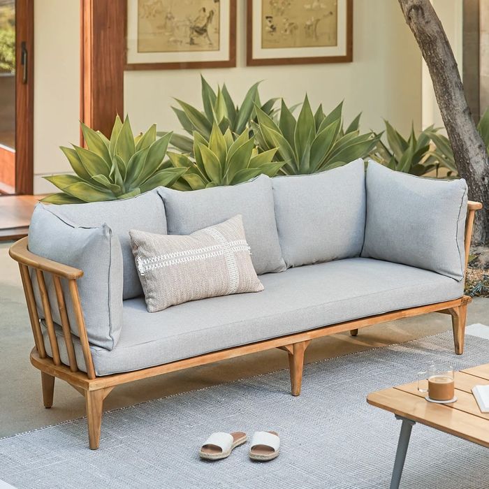 A wooden outdoor sofa with plush cushions and a decorative pillow, set amongst plants on a patio, with slippers and a table in the foreground.